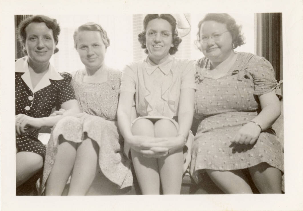 Black and white photograph of four women all sitting near each other. They all wear dresses.